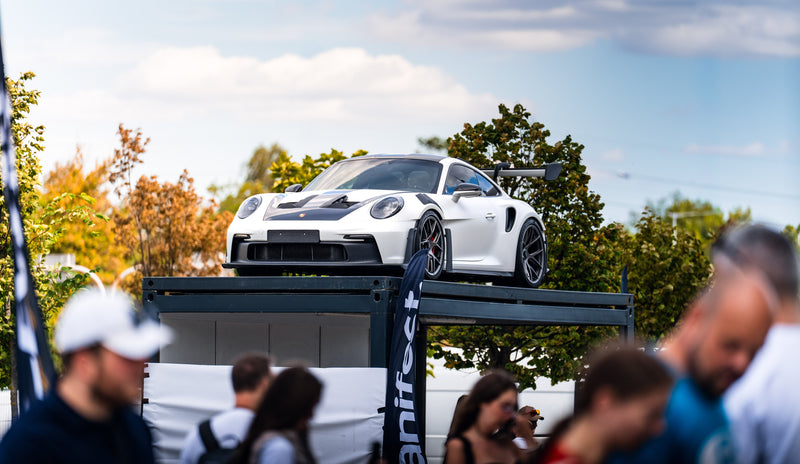White sports car on a lift with people in the foreground and trees in the background